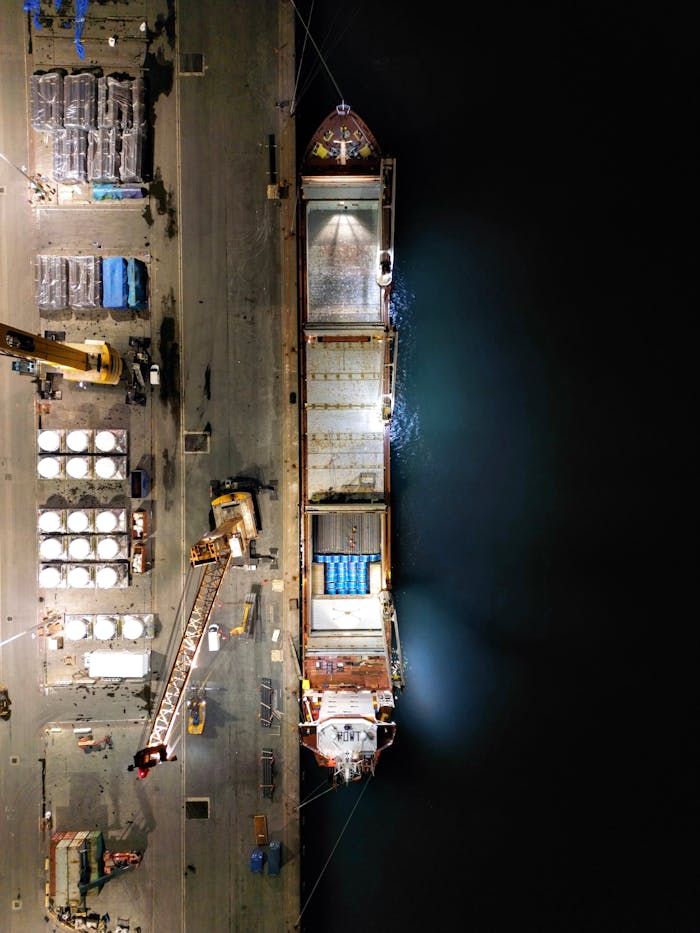 Top-down drone shot of an illuminated cargo ship docked at a bustling nighttime port.