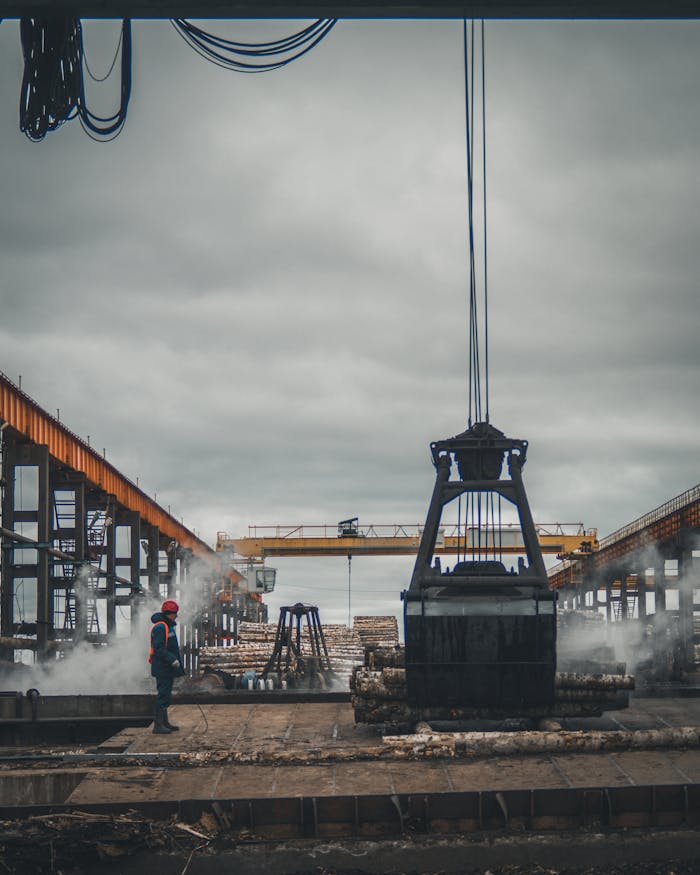 A large crane moving cargo at an industrial dock with a worker overseeing operations under cloudy skies.